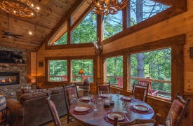Spacious Great Room With Plenty of Natural Light Aska Escape Lodge | Blue Ridge Cabins | Cabin Rentals of Georgia | Dining Area for 6 guests in open floor plan with living area and fireplace with cathedral ceiling, antler chandeliers, and a wall of windows looking at the wooded view
