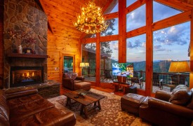 Riverview Lodge | Cabin Rentals of Georgia | Grand lodge living area with floor-to-ceiling windows at dusk with antler chandelier and fire in the stone fireplace, leather furnishings